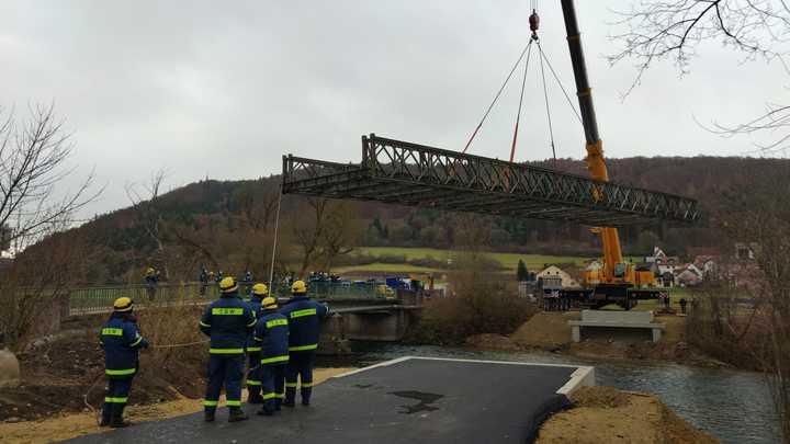 Ein Autokran hebt die Bailey-Brücke auf die vorgefertigten Fundamente. Im...